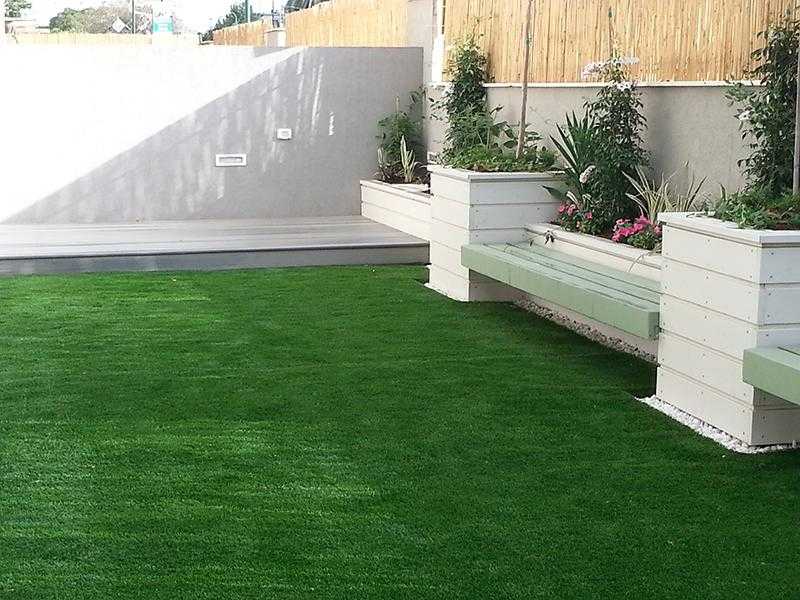 דשא סינטתי במת עץ מוגבהת ספסלים ואדניות מובנות בגינת חצר