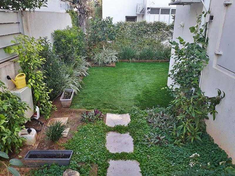 שילוב של אבני מדרך דשא עצים אדניות וצמחייה מעורבת בגינת חצר במרכז הארץ